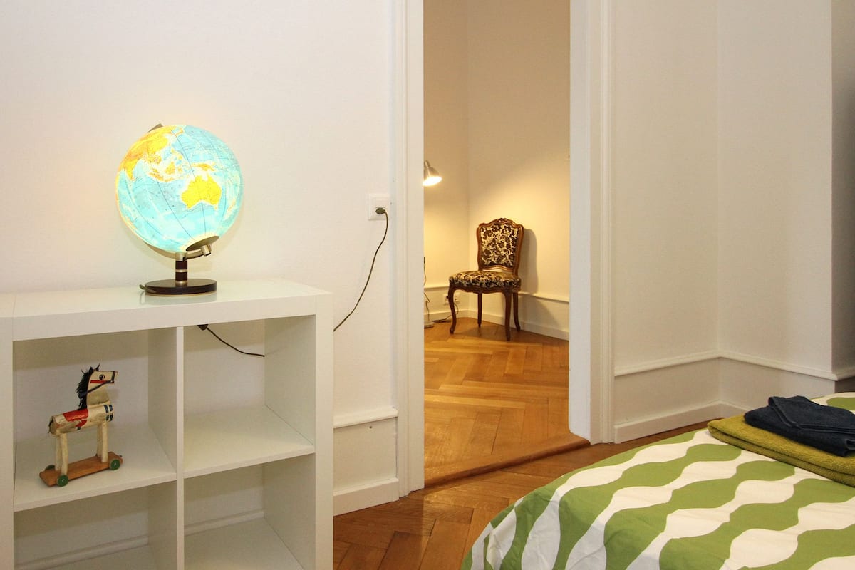 Une pièce avec un globe terrestre posé sur une étagère blanche, une chaise et un lit recouvert d’une couvre-lit à rayures vertes et blanches.