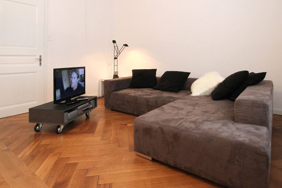 Un salon avec un canapé d’angle, un téléviseur posé sur un meuble à roulettes et une lampe.