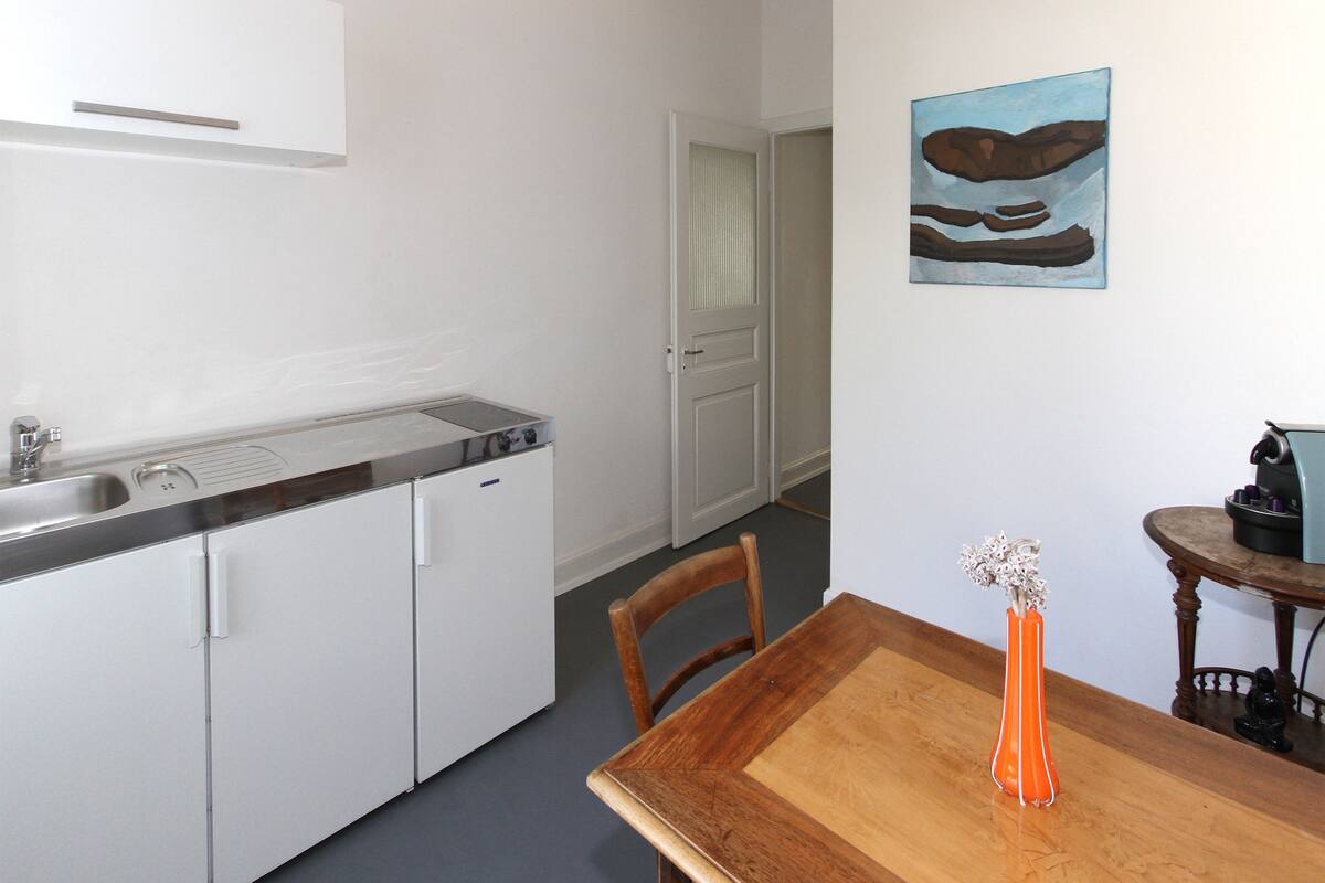 Une cuisine compacte équipée d’un évier, d’une cuisinière et de meubles blancs. On y trouve une table à manger, un vase et un tableau accroché au mur.