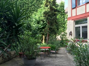 A courtyard with a red table and chairs, surrounded by potted plants and a tall tree.
