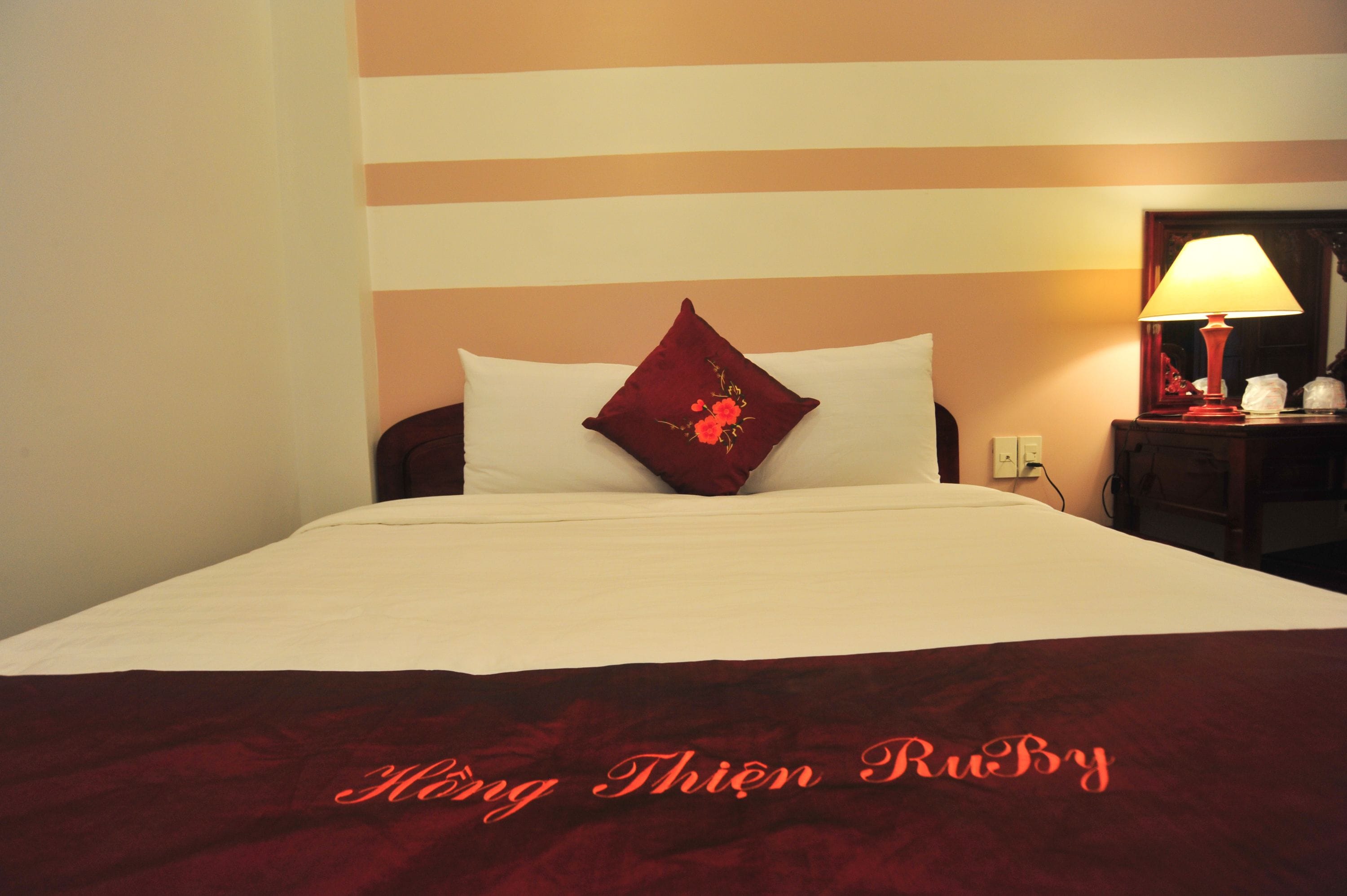 A hotel room with a bed, a red throw blanket, and a lamp on a bedside table.