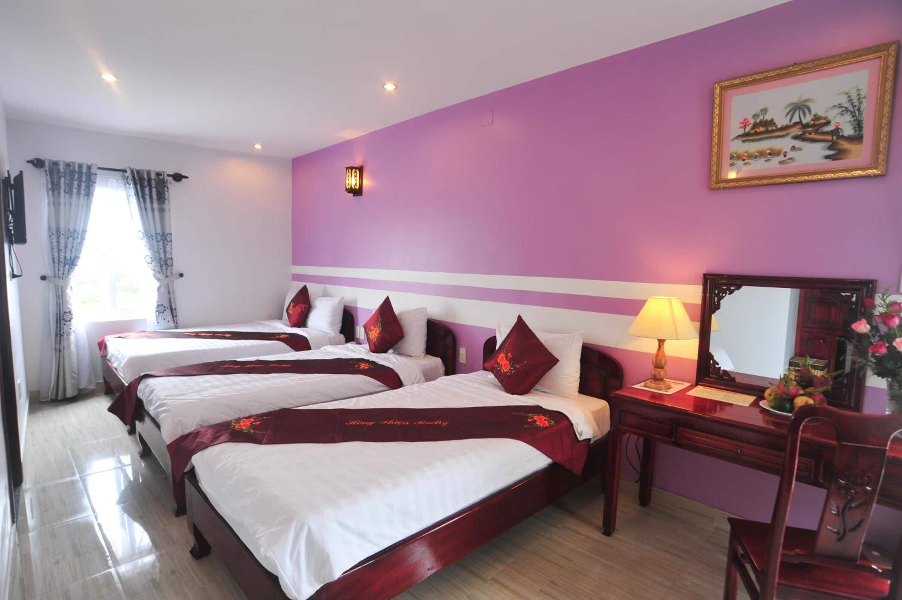 Habitación de hotel con tres camas, un escritorio con lámpara, un espejo y un cuadro en la pared.