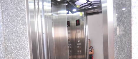 Interior de ascensor con paredes metálicas, puerta de vidrio y alfombra roja.