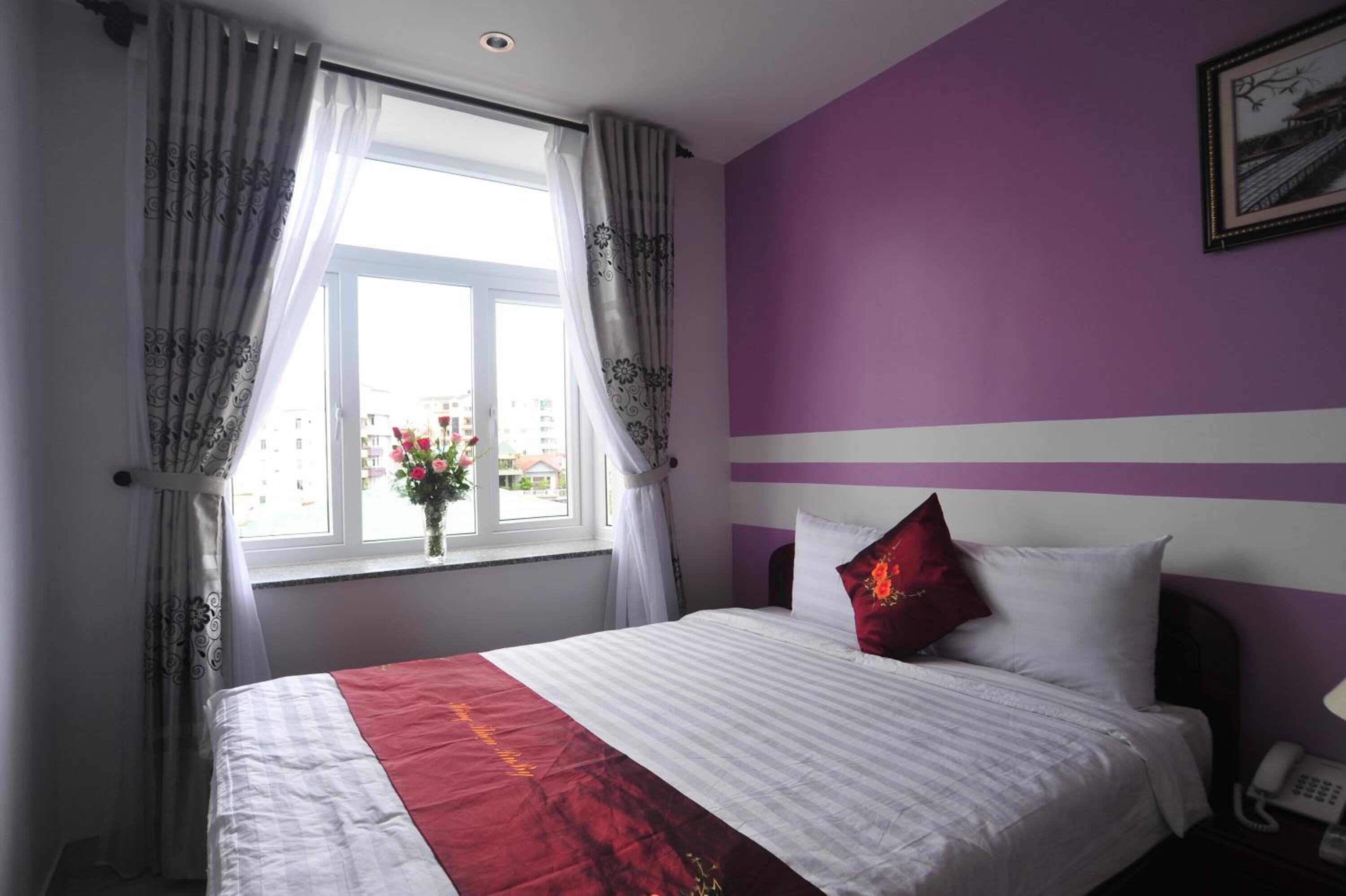 A hotel room with a bed, a window with curtains, a vase with flowers, and a wall with a picture.