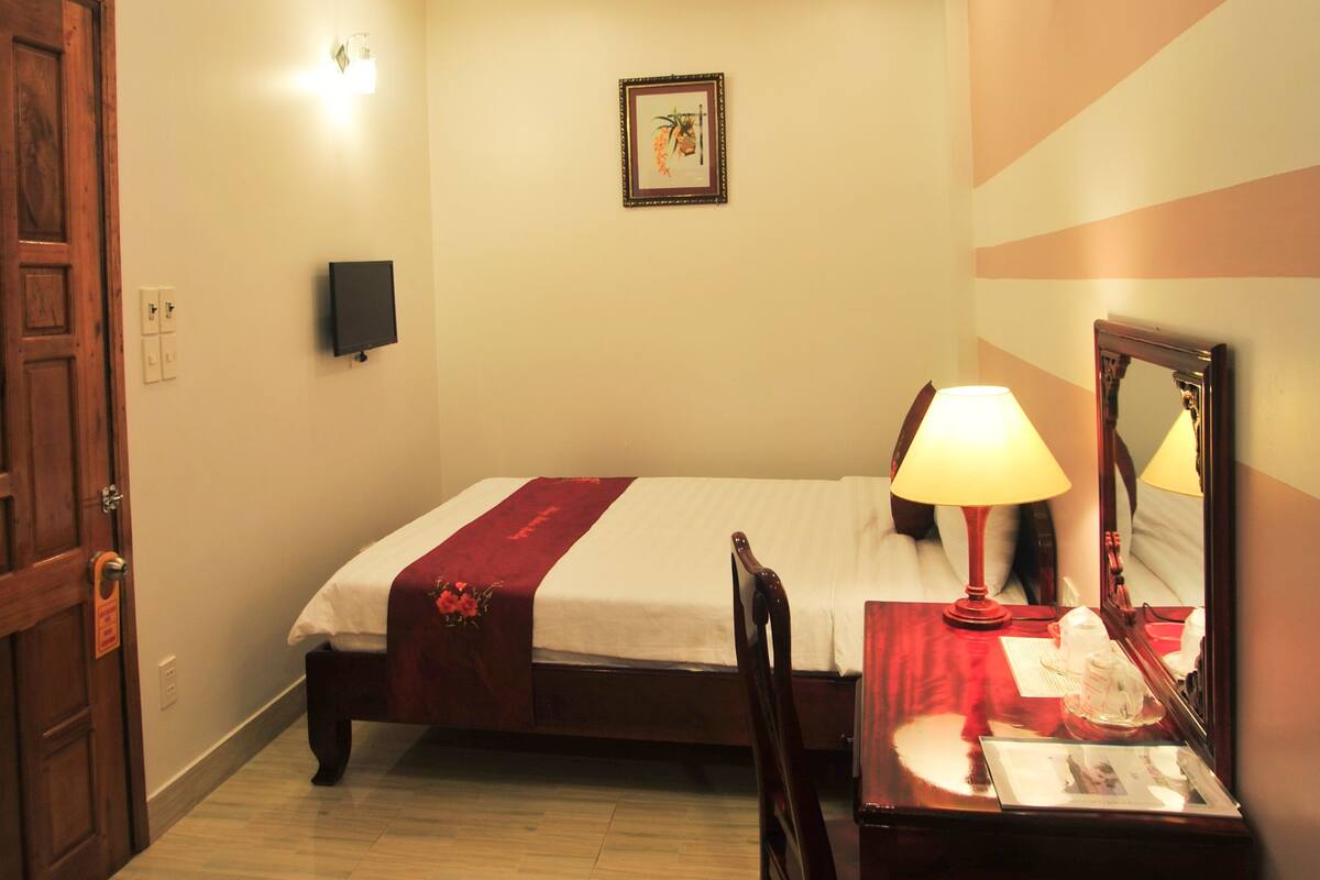 Habitación de hotel con cama, mesita de noche con lámpara, una silla, un espejo y televisor montado en la pared.