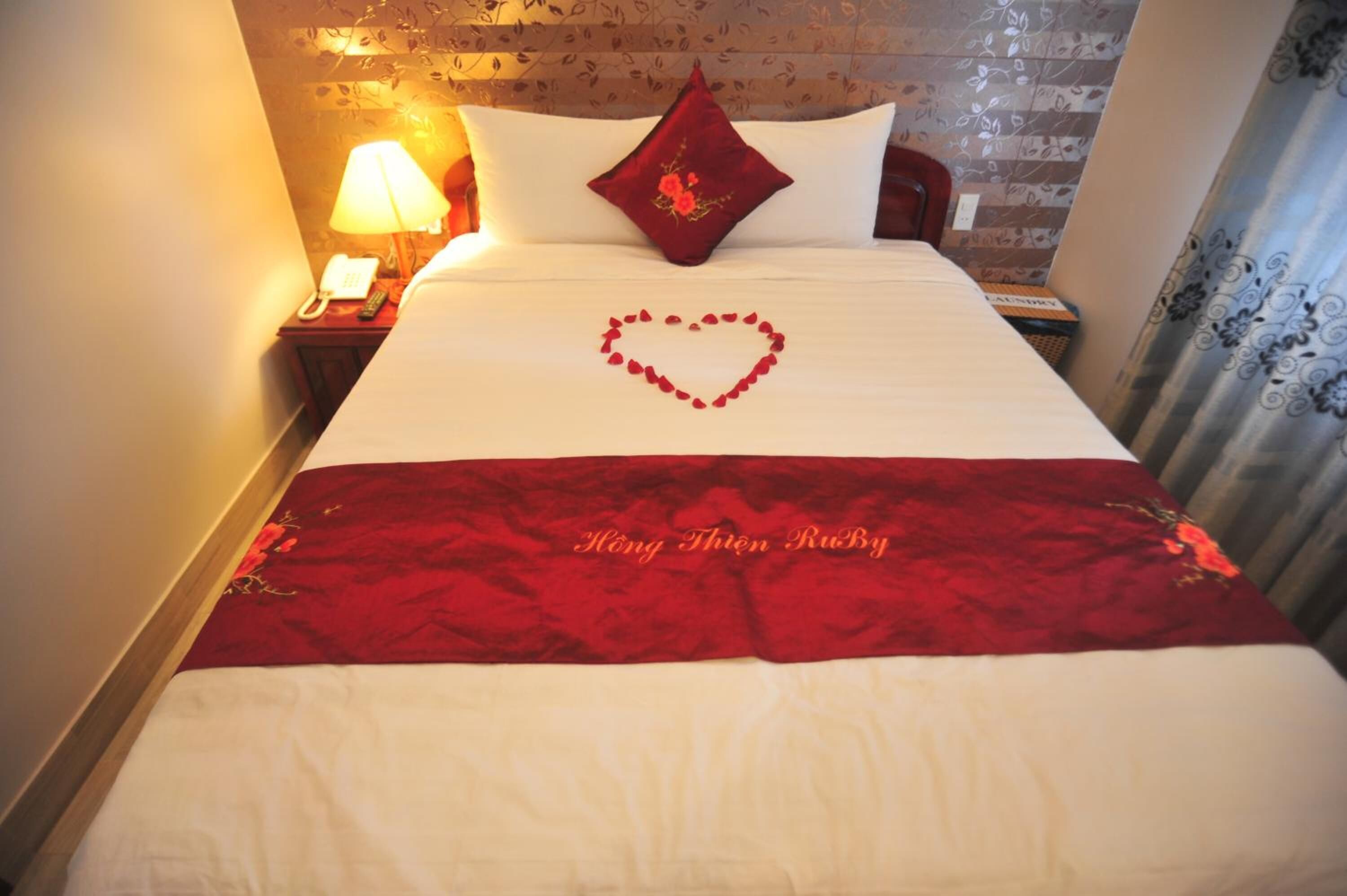 A hotel room with a neatly made bed, a heart-shaped decoration on the bed, and a red throw blanket with text.