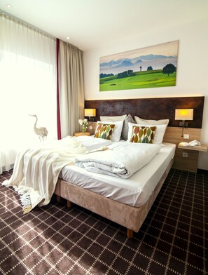 A hotel room with a large bed, two bedside tables, a nightstand, a wall-mounted lamp, a painting of a landscape, and a window with curtains. - Business Class Hotel Ebersberg (Ebersberg)