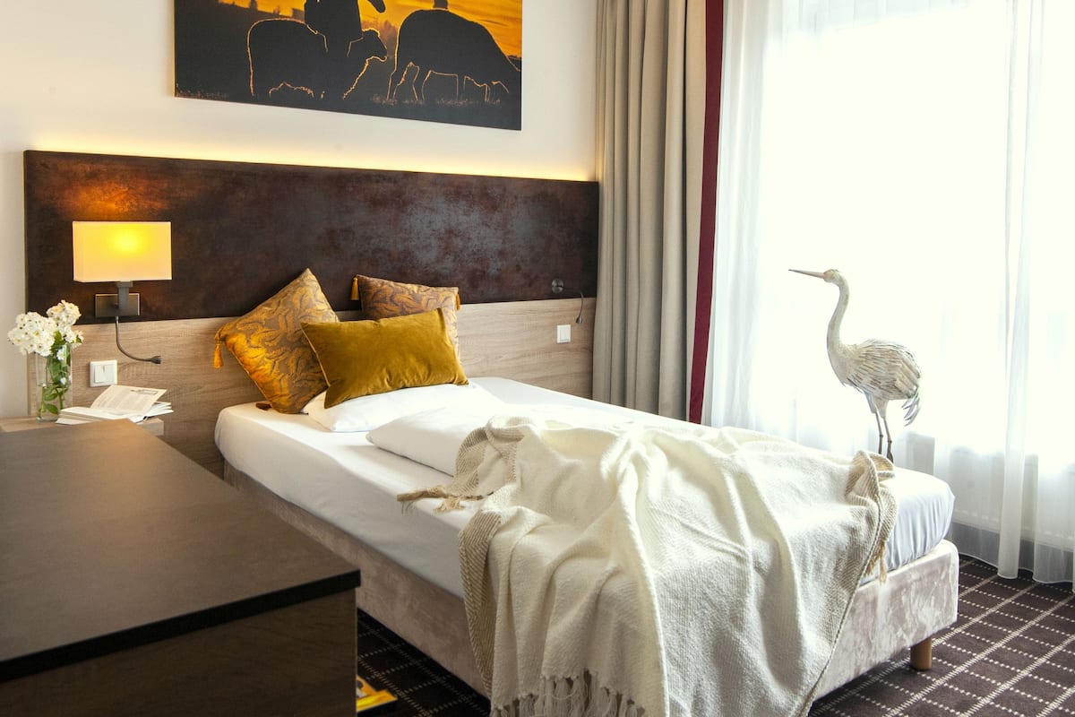A hotel room with a large bed, a nightstand, a lamp, a painting, and a window with curtains.