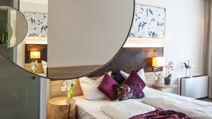 A hotel room with a large mirror, a bed with purple pillows, a bedside table with a lamp, and a wall-mounted artwork. - Business Class Hotel Ebersberg (Ebersberg)