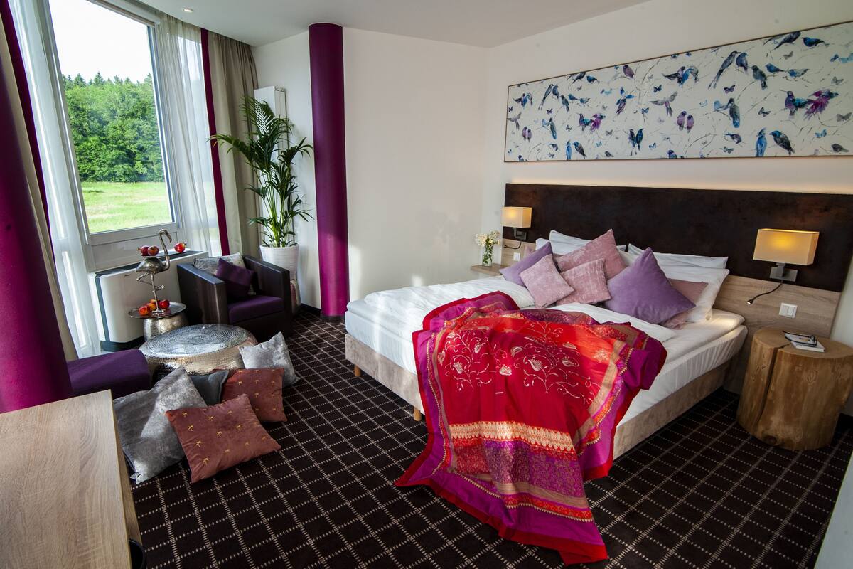 A hotel room with a bed, a sofa, a small table, a potted plant, and a view of the outdoors.