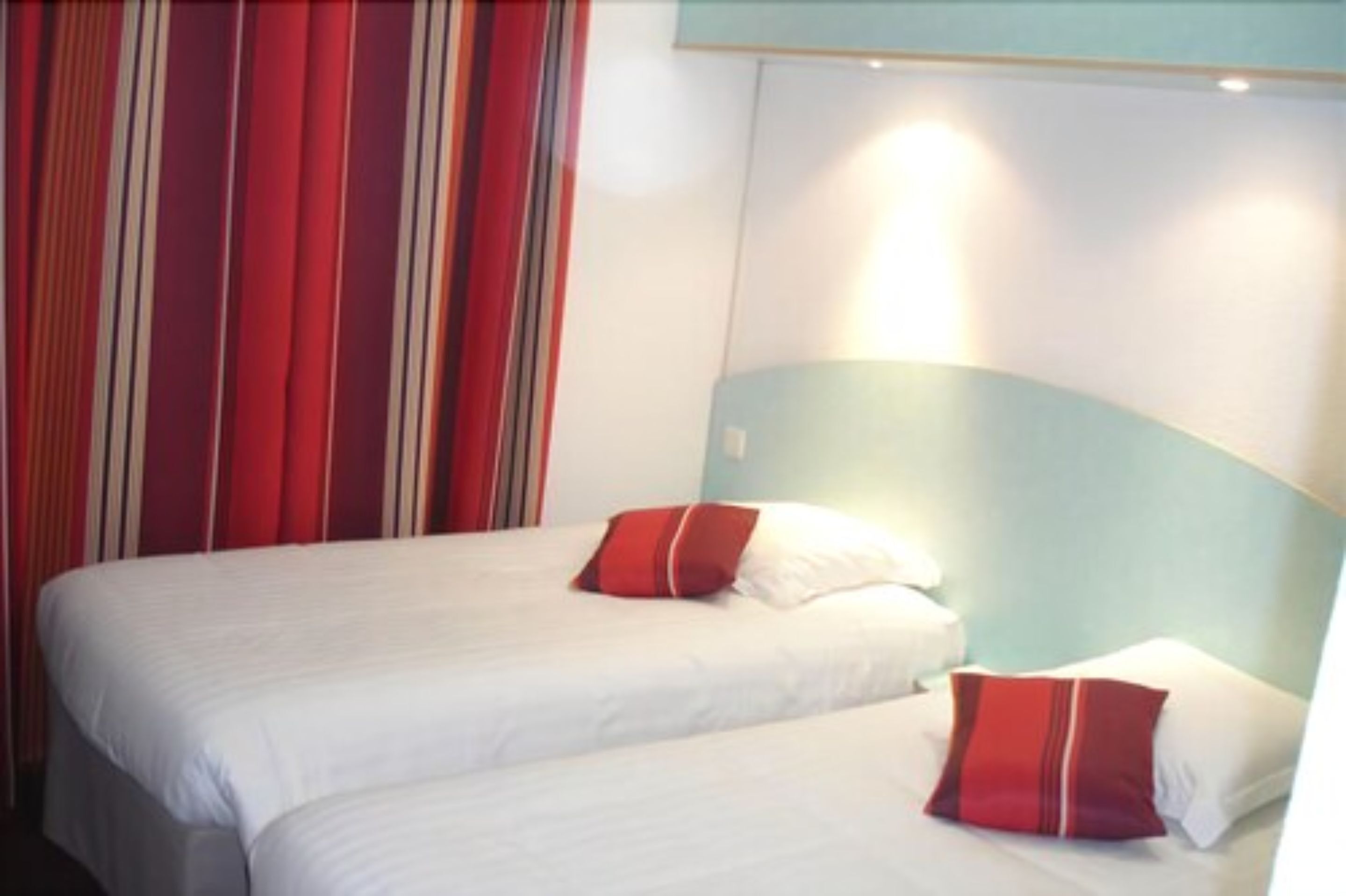 Hotellihuone, jossa on kaksi sänkyä. Kummassakin sängyssä on valkoiset lakanat ja punaiset tyynyt, ja taustalla on raidallinen verho.