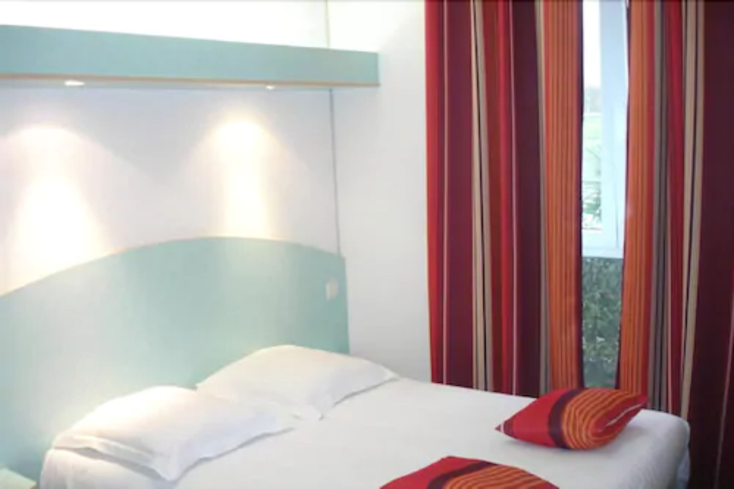 Une chambre d’hôtel avec un lit, des oreillers blancs, un oreiller à rayures rouges et oranges, et une fenêtre avec des rideaux.