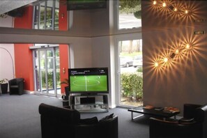 A modern lobby with a large screen displaying a soccer game, comfortable seating, and a view of the outdoors. - Agape Hotel (Bessines)