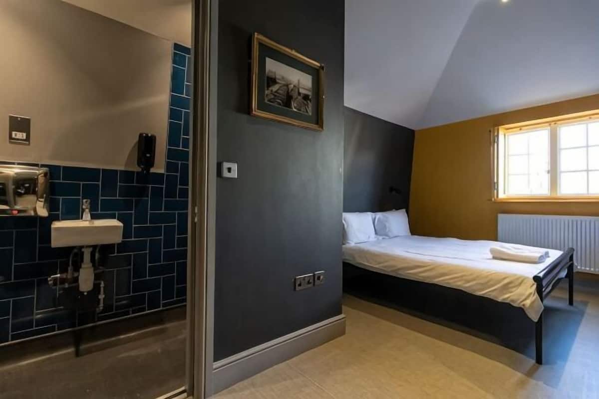 A bedroom with a bed, a window, a radiator, and a bathroom with a sink and mirror.