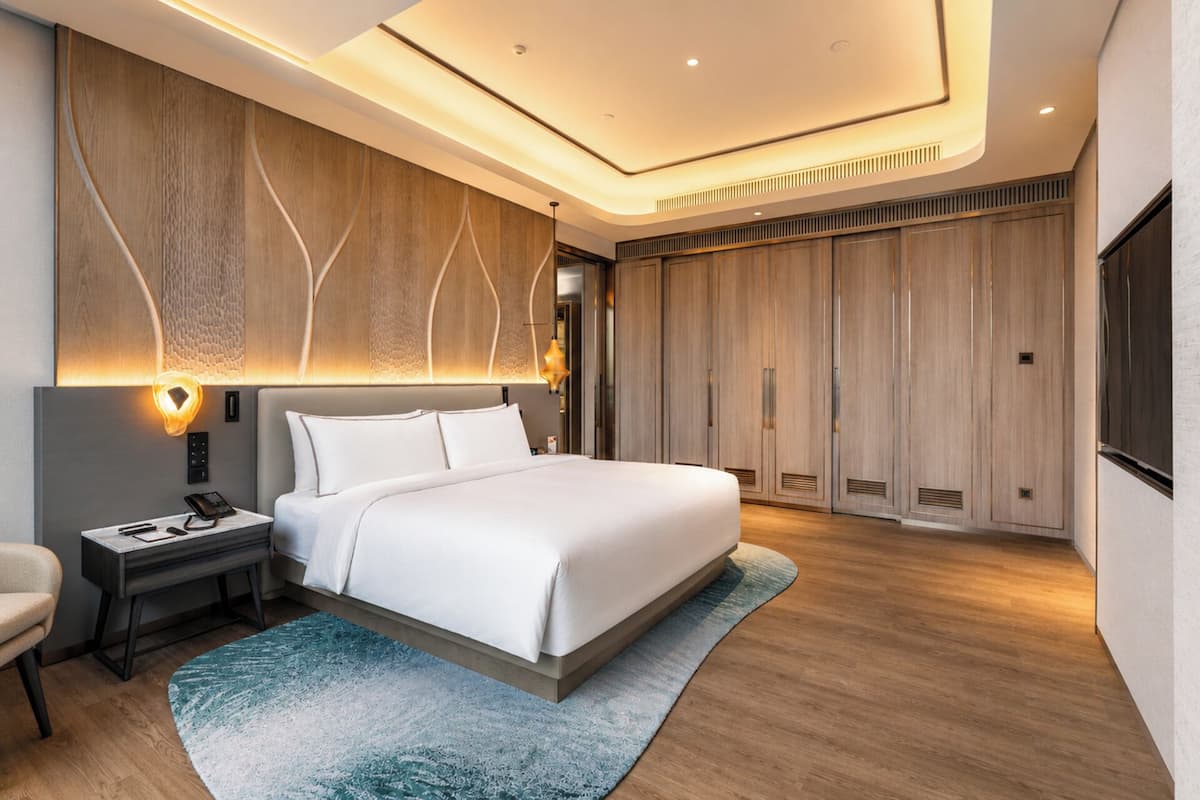 A modern hotel room with a large bed, a desk with a telephone, a chair, and a wardrobe.