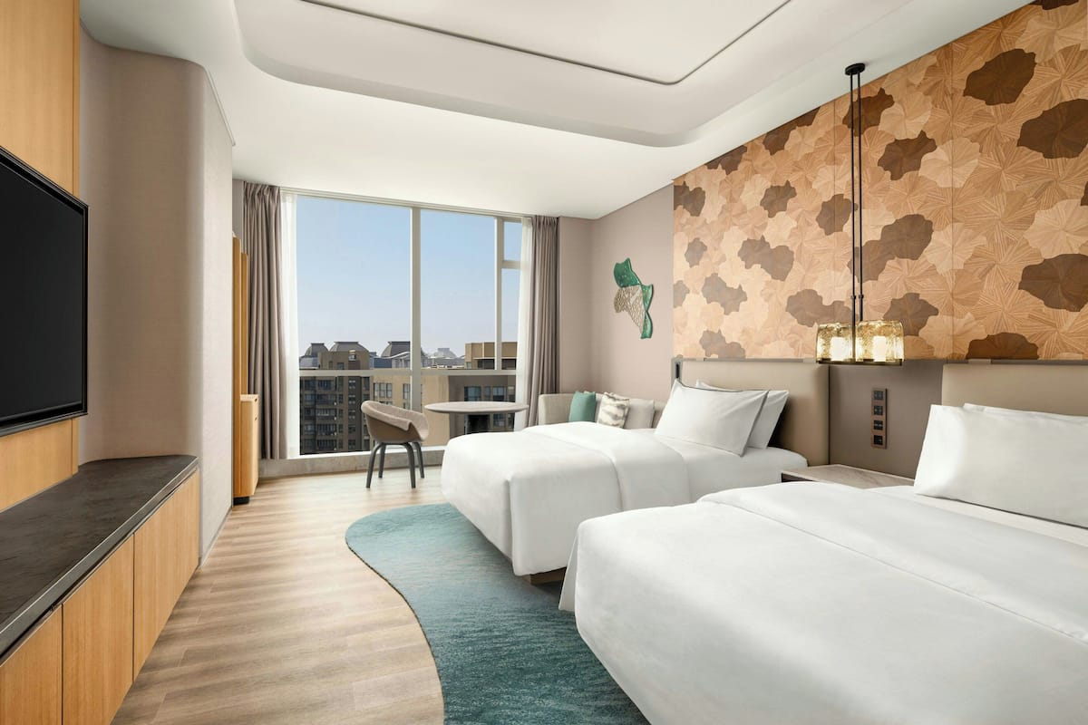 A hotel room with two beds, a TV, a desk, and a large window with a city view.