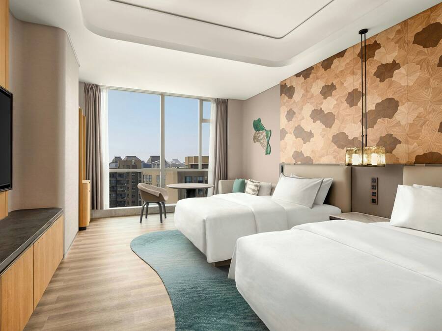 Quarto de hotel com duas camas, televisão, escrivaninha e uma janela grande com vista para a cidade.