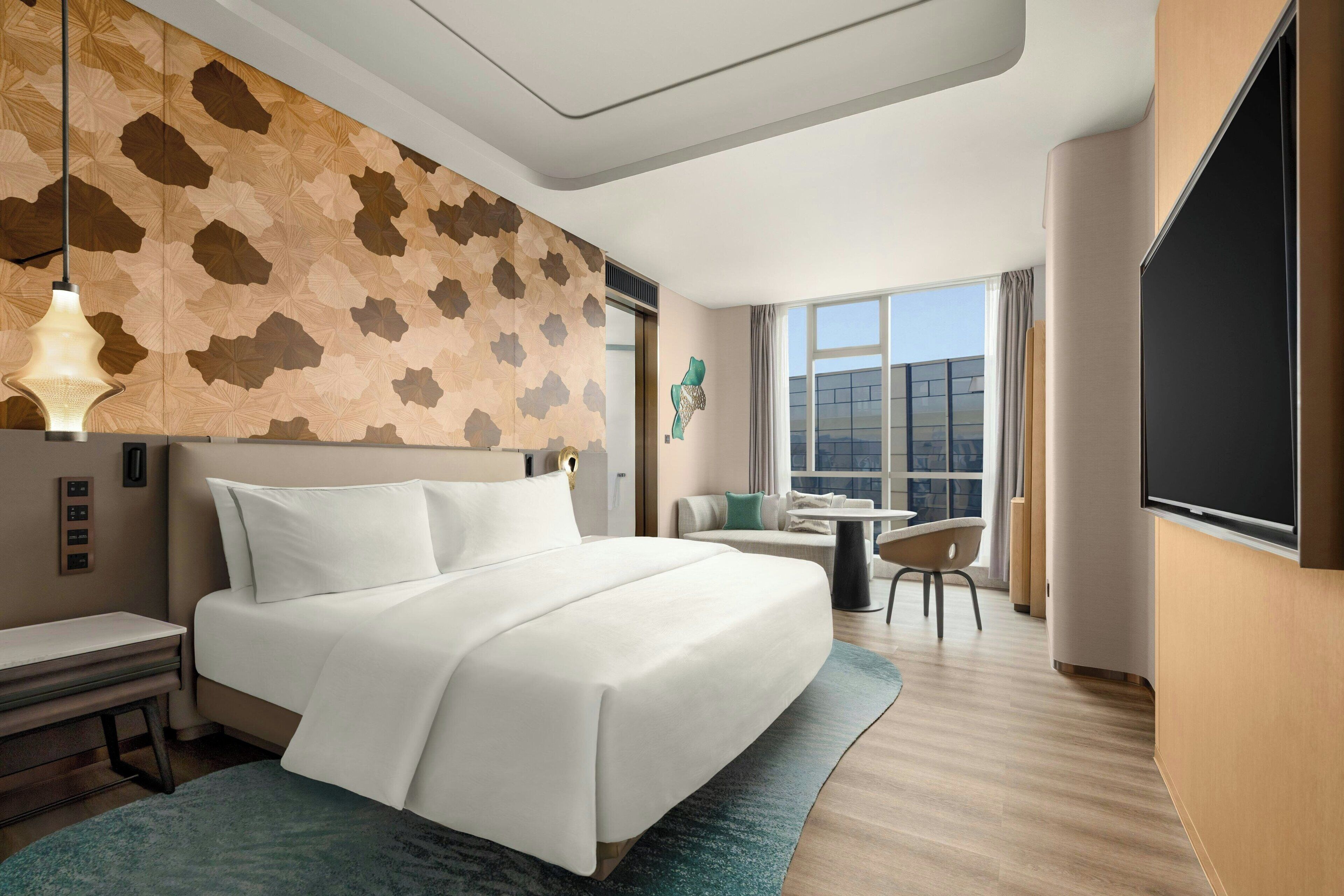 Habitación de hotel con una cama grande, un escritorio, una silla, una mesita, televisión y ventana con vista a la ciudad.