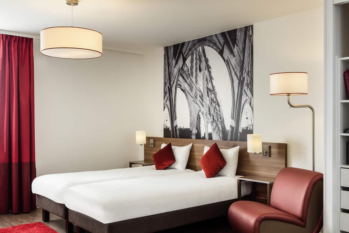 Een hotelkamer met een bed, twee nachtkastjes, een rode stoel, een rood kleed en een groot zwart-wit kunstwerk aan de muur.