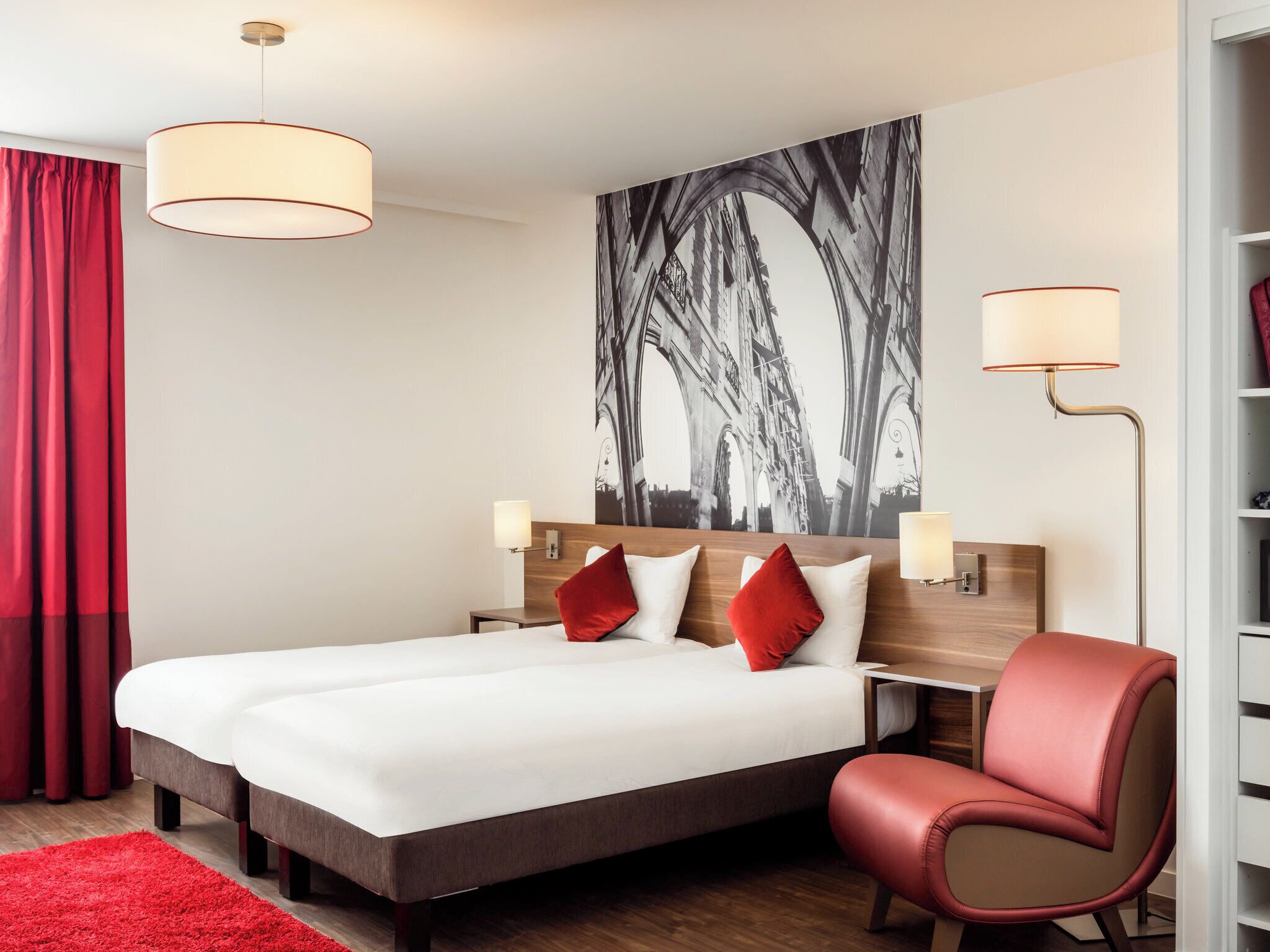 A hotel room with a large bed, two bedside tables, a red chair, a red rug, and a large black-and-white artwork on the wall.