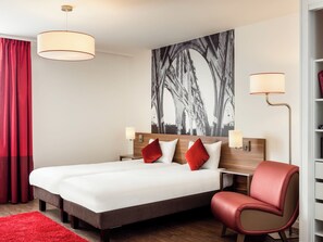 Een hotelkamer met een groot bed, twee nachtkastjes, een rode stoel, een rood vloerkleed en een groot zwart-wit kunstwerk aan de muur.