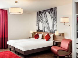 Quarto de hotel com uma cama grande, duas mesas de cabeceira, uma cadeira vermelha, um tapete vermelho e uma grande obra de arte em preto e branco na parede.