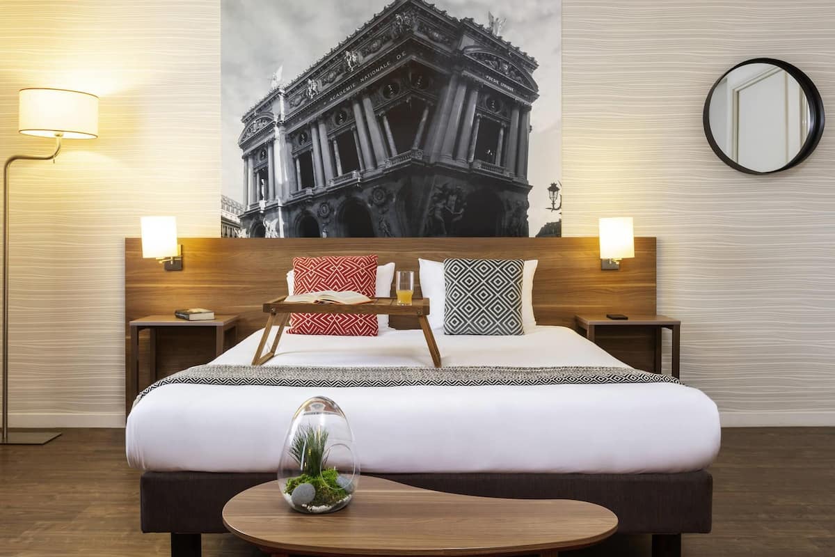 Ein Hotelzimmer mit einem groĂen Bett, einem Nachttisch, einer Lampe, einem Spiegel und einem an der Wand hĂ€ngenden Bild eines GebĂ€udes.