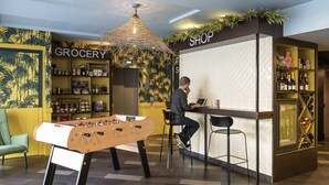 Un espace d’accueil avec un baby-foot, un homme qui travaille derrière un comptoir portant l’inscription « MAGASIN », et un rayon « ÉPICERIE » avec des étagères remplies de produits.