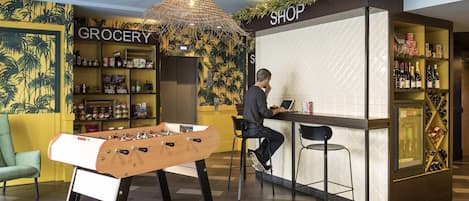 Un área de recepción con una mesa de fútbol de mesa, un hombre trabajando en un mostrador con el rótulo 'SHOP' y una sección de 'GROCERY' con estantes llenos de productos.