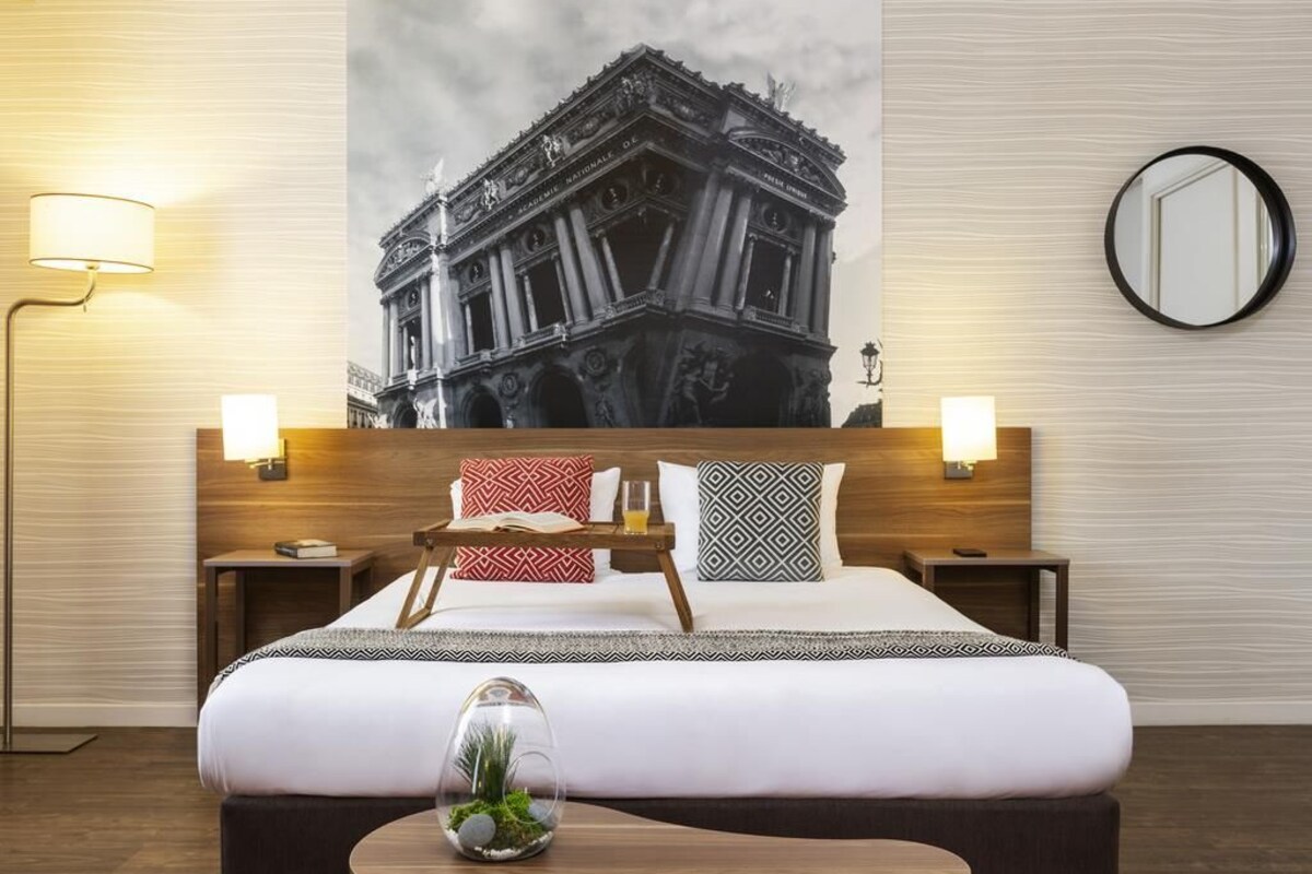 Een hotelkamer met een groot bed, twee nachtkastjes, een ronde spiegel en een aan de muur hangend schilderij van een historisch gebouw.