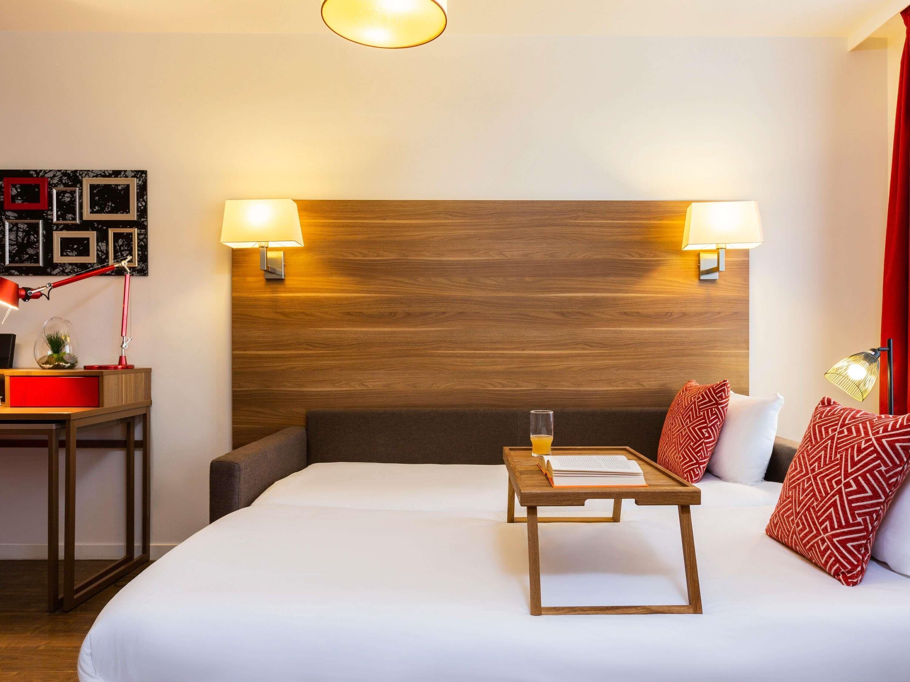 A hotel room with a wooden headboard, two bedside lamps, a bedside table with a book and a glass of orange juice, and a red and white patterned pillow.