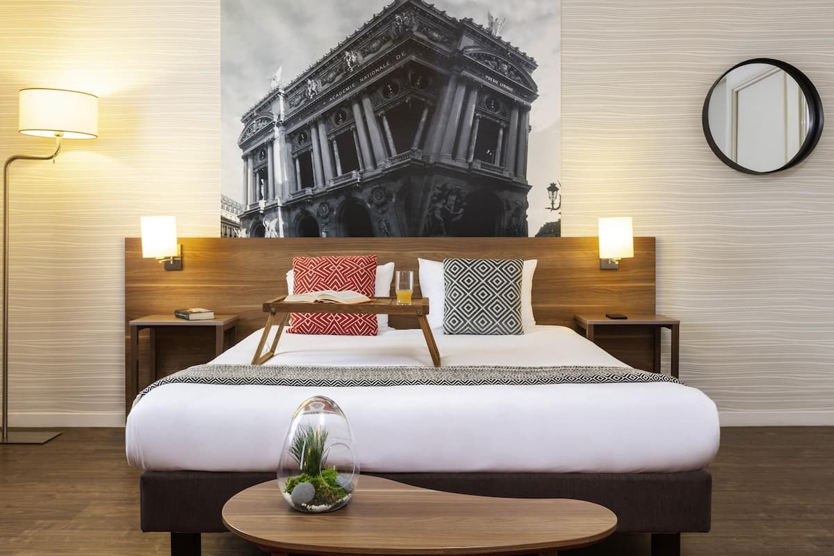 Een hotelkamer met een groot bed, twee nachtkastjes, een spiegel, een wandlamp en een decoratief object op het nachtkastje.