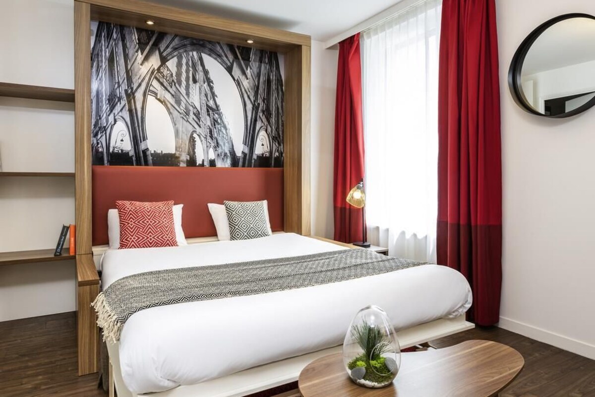 Een hotelkamer met een bed, een hoofdeinde met een stadsgezichtsmural, een ronde spiegel en een tafeltje met een plant.