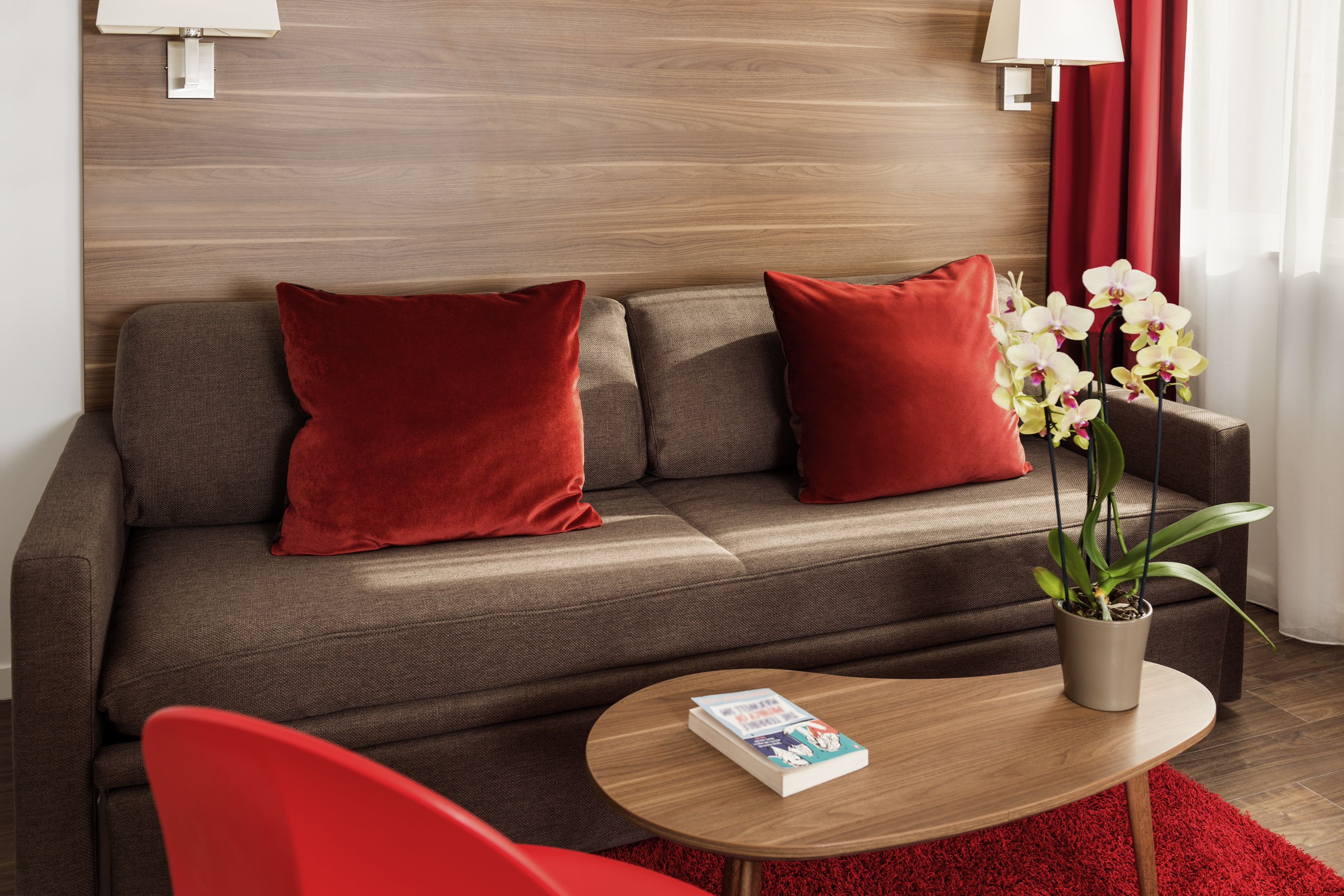 Een moderne woonkamer met een grijze bank, rode kussens, een klein rond tafeltje met een boek en een potorkidee.