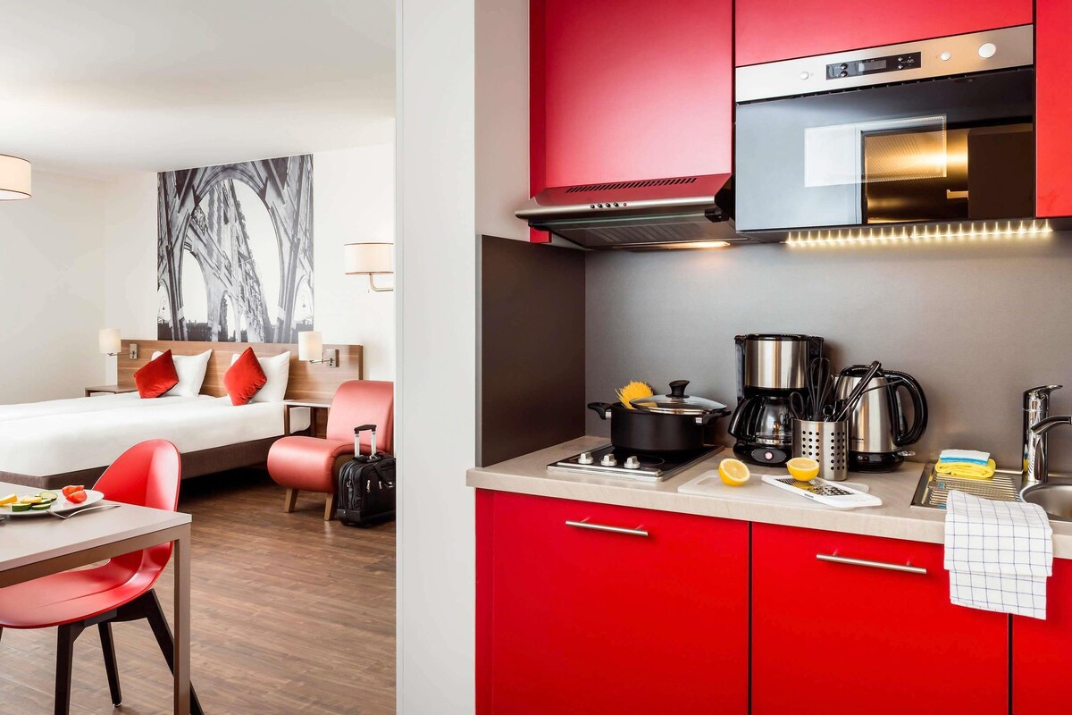 Een hotelkamer met een bed, een eettafel met stoelen, een kitchenette met rode kastjes en een groot raam.
