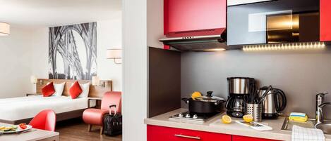 Een hotelkamer met een bed, een eettafel met stoelen, een kitchenette met rode kastjes en een groot raam.