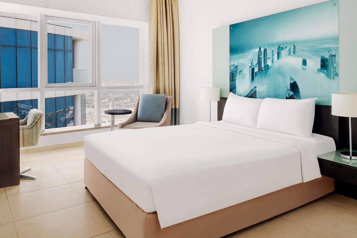 Ein Hotelzimmer mit einem großen Bett, einem Sessel, einem Schreibtisch und einem Fenster mit Blick auf die Stadt.