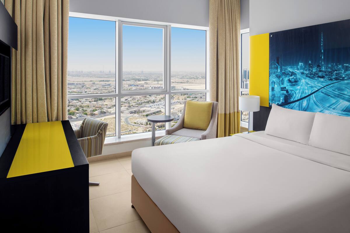 Ein Hotelzimmer mit einem großen Bett, einem Sessel, einem kleinen Tisch und Blick auf eine Stadtlandschaft durch große Fenster.