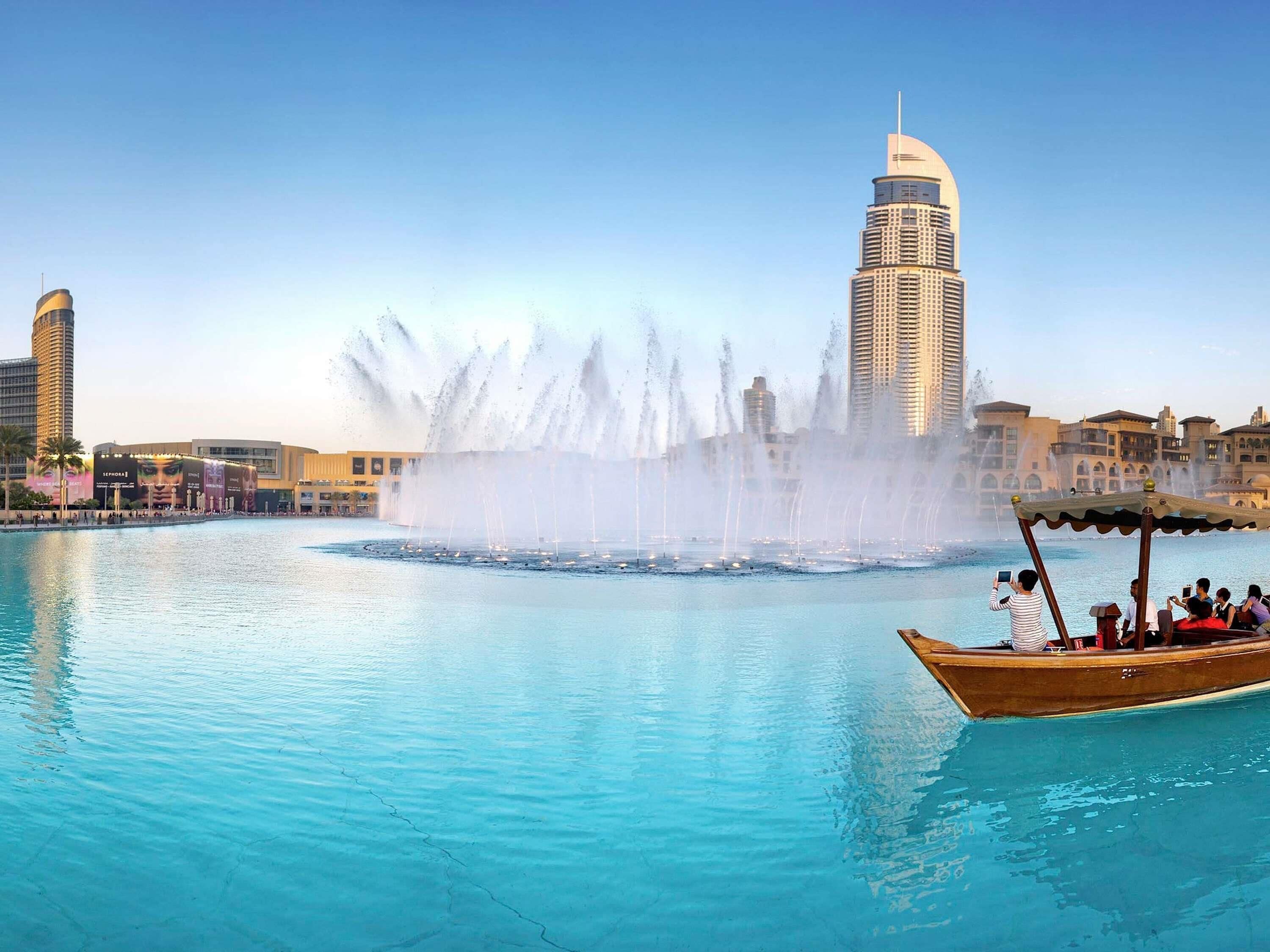 A waterfront view with a large fountain, a tall building, and a boat with people.