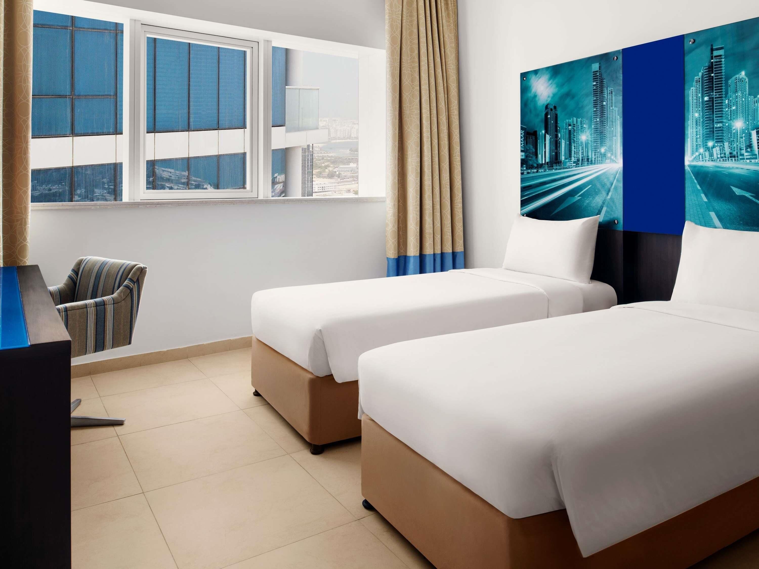 Une chambre d’hôtel avec deux lits, une chaise, un bureau et une grande fenêtre donnant sur la ville.