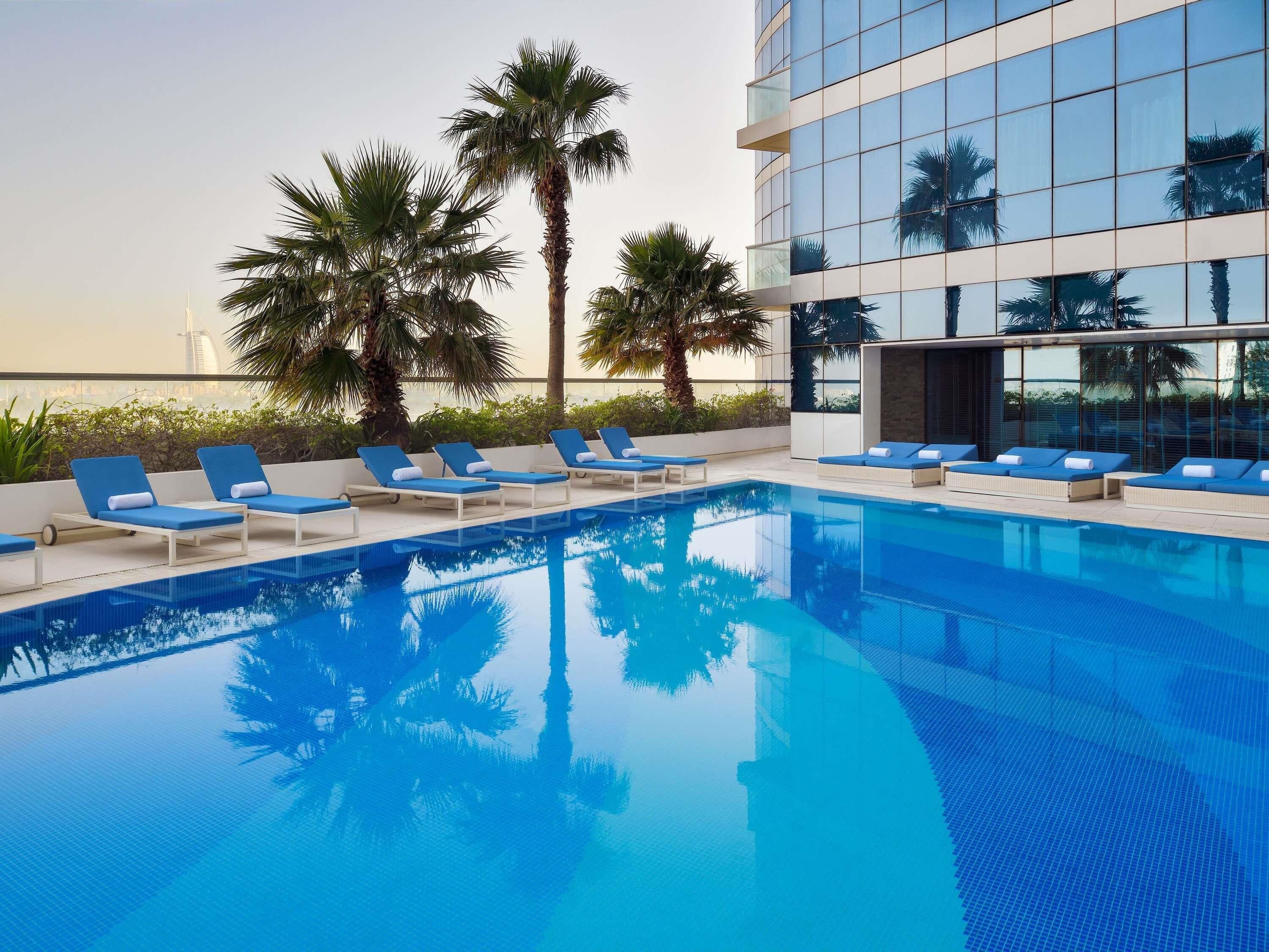 A modern hotel pool with blue lounge chairs, palm trees, and a glass building in the background.