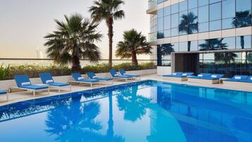 A modern hotel pool with blue lounge chairs, palm trees, and a glass building in the background.