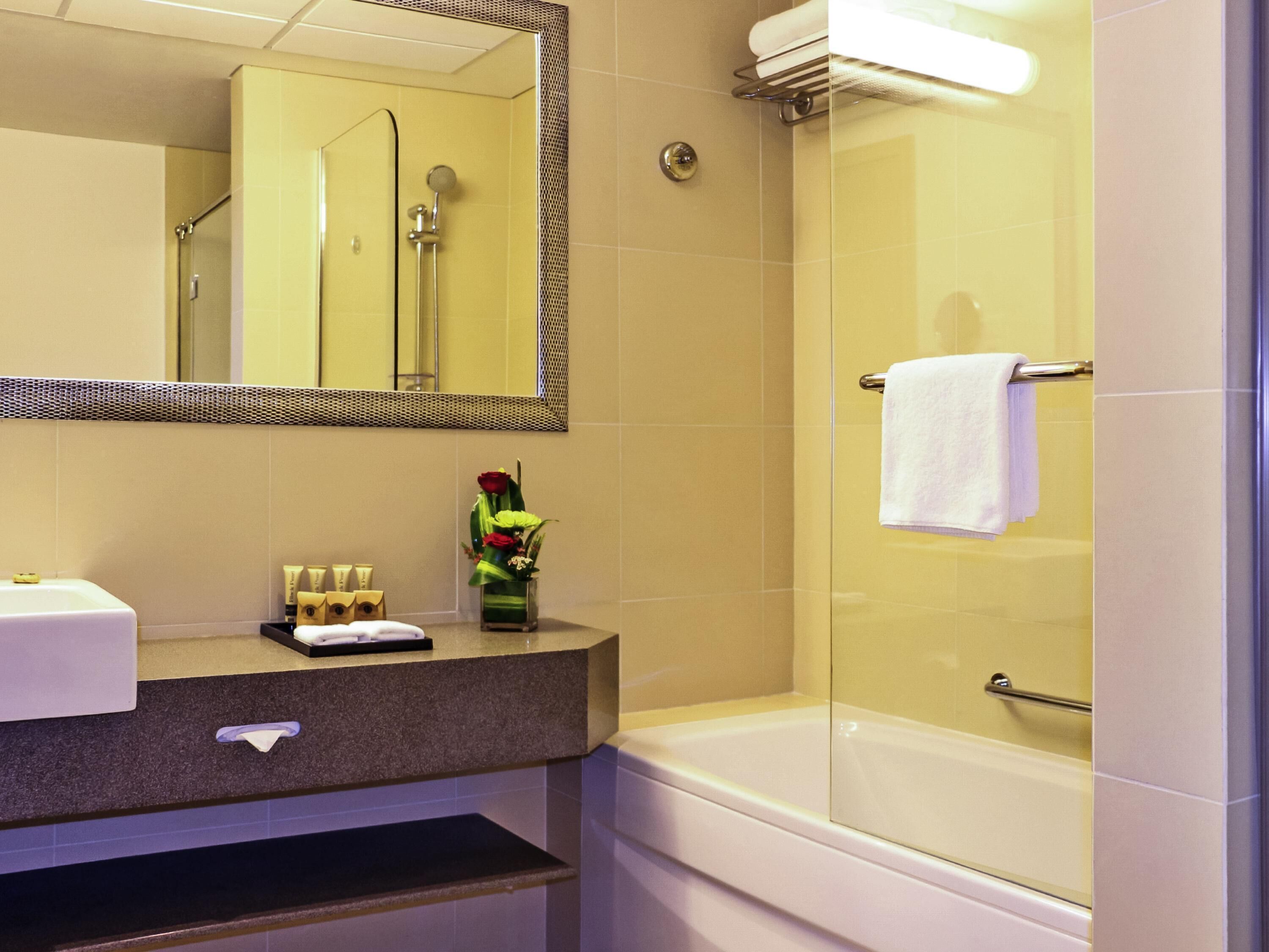 A bathroom with a sink, mirror, toiletries, a vase with flowers, and a bathtub.