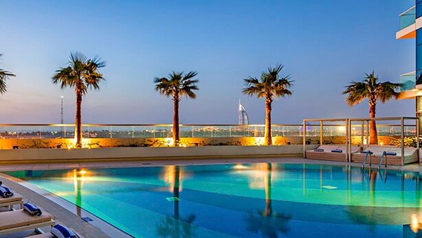 A swimming pool with lounge chairs and palm trees, overlooking a cityscape with a tower.