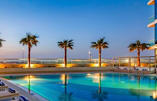 Una piscina con sillas de descanso y palmeras, con vistas a un paisaje urbano que incluye una torre.