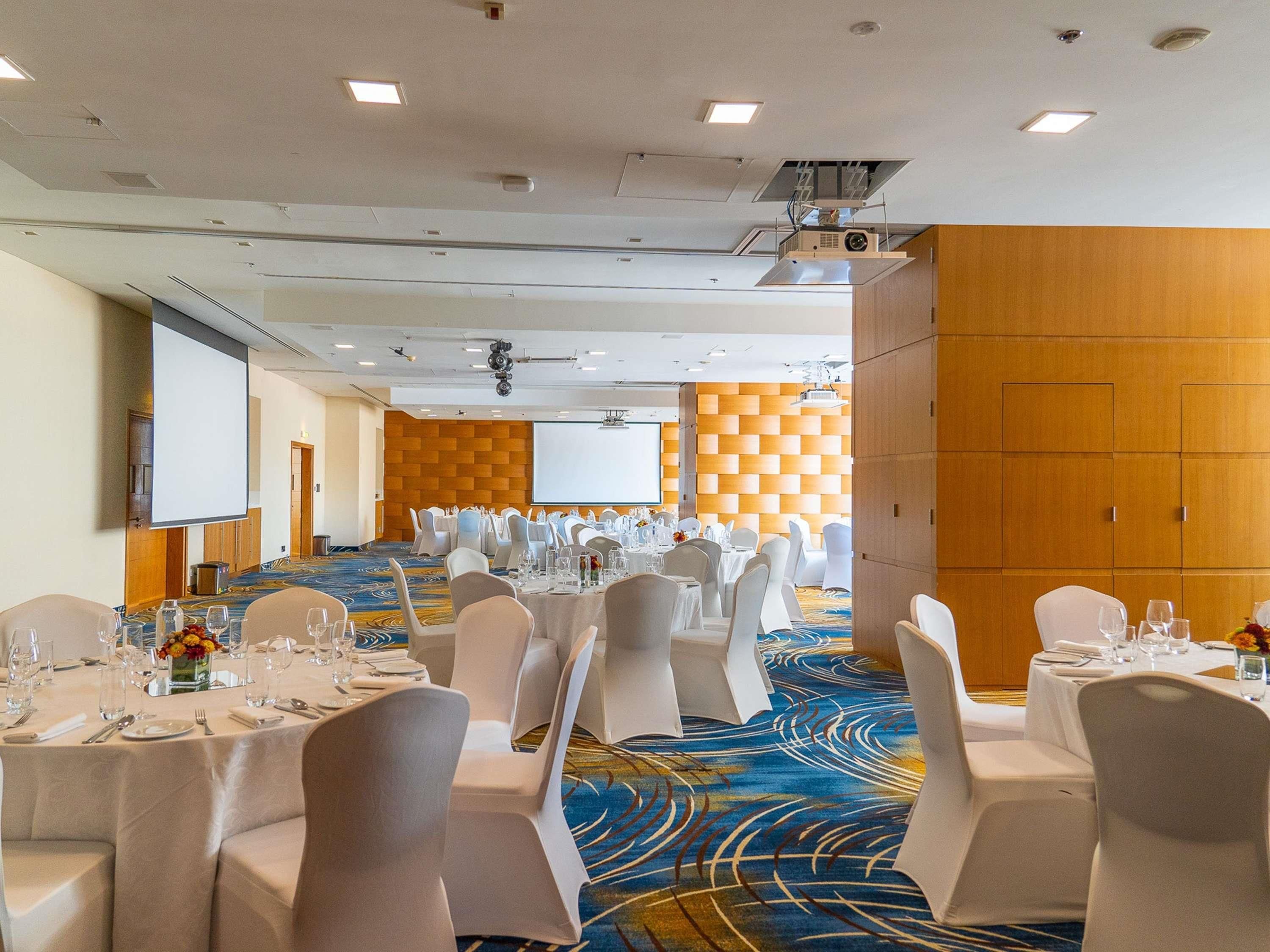 Un salón de conferencias con varias mesas dispuestas para un evento formal, equipado con proyectores y una pantalla grande.