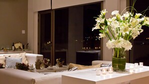 A modern dining area with a white table set for two, a vase of white flowers, and a view of the city at night through large windows. - Brizo Neuquen (Neuquén)