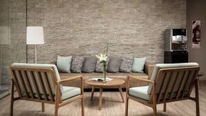 A modern living room with a brick-patterned wall, a sofa with grey cushions, two wooden chairs, a round table with a vase of flowers, and a coffee machine. - Brizo Neuquen (Neuquén)