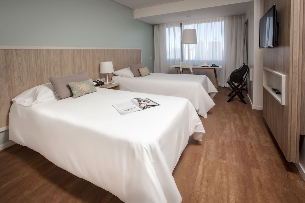 Quarto de hotel com duas camas, uma escrivaninha, uma cadeira, televisão e janela com cortinas.