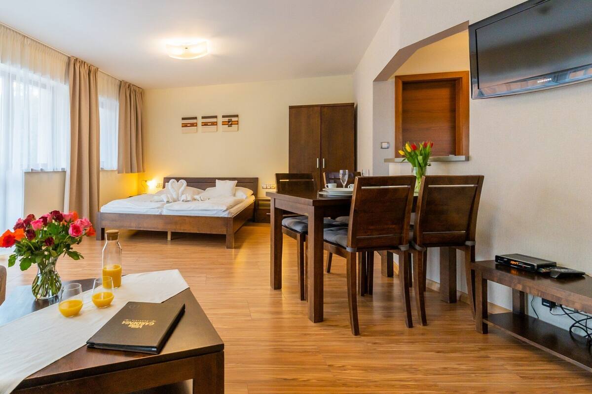 A hotel room with a bed, a desk, a dining table with chairs, a TV mounted on the wall, and a window with curtains.